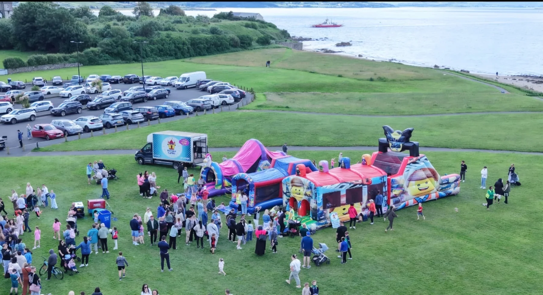 Aerial view of a Druminor Castles setup at a coastal community event, with several inflatables and a large crowd